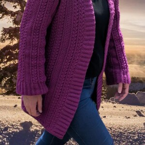 May include: A long, open-front, purple crochet cardigan with a textured stitch pattern. The cardigan is knee-length and has long sleeves. The person is wearing a black shirt and blue jeans. The background is a blurred outdoor scene.