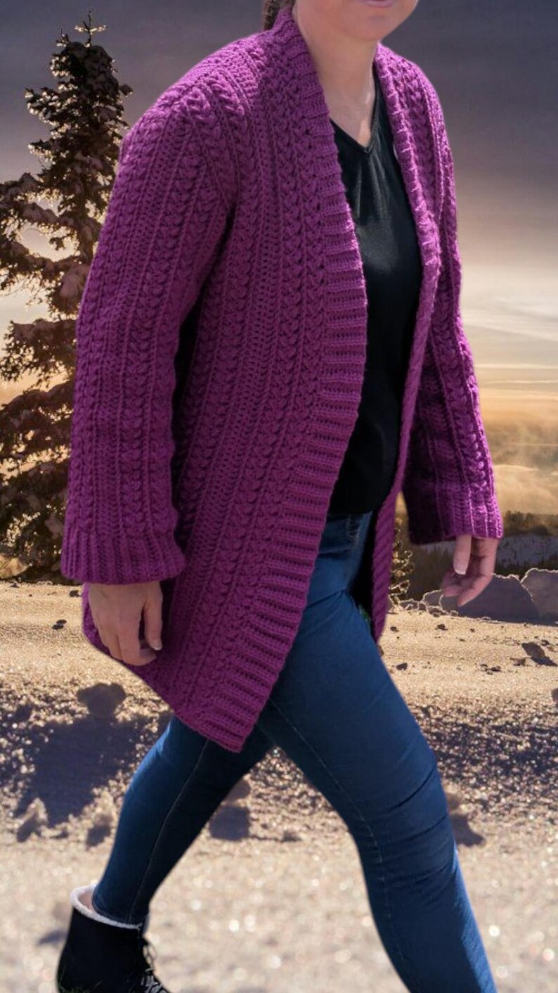 May include: A long, open-front, purple crochet cardigan with a textured stitch pattern. The cardigan is knee-length and has long sleeves. The person is wearing a black shirt and blue jeans. The background is a blurred outdoor scene.