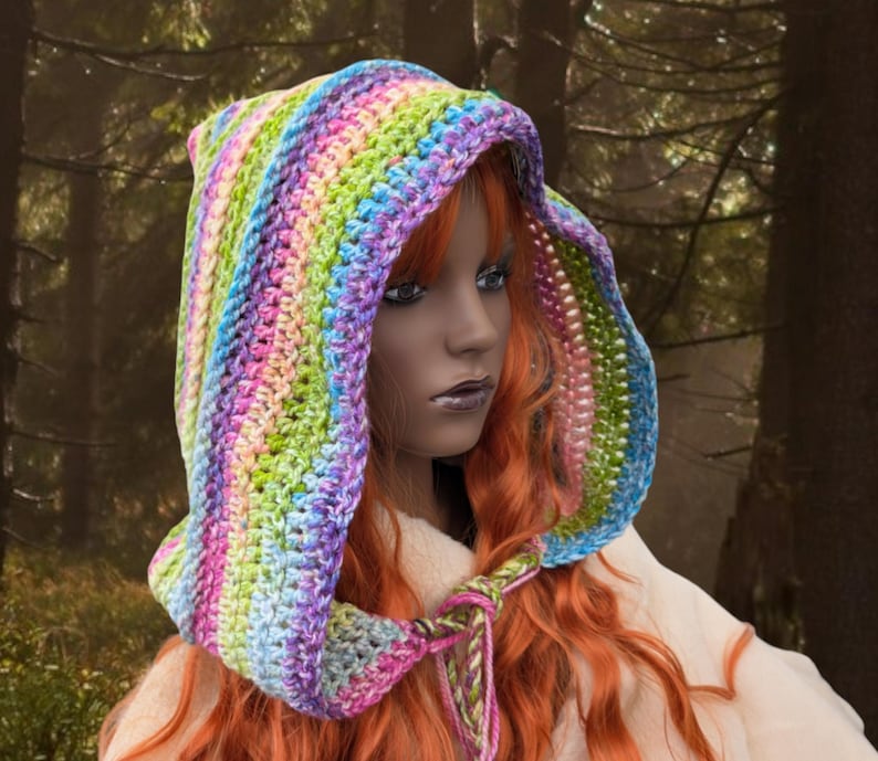 May include: A colourful crocheted hooded scarf with rainbow stripes in shades of pink, purple, green, blue, and white. The scarf is worn by a person with long red hair.