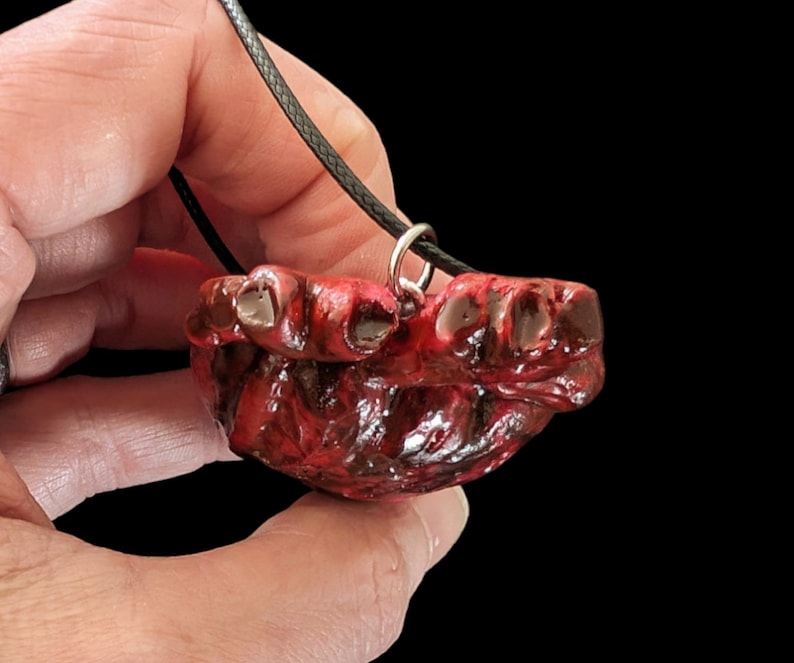 Op de afbeelding: Een rode en zwarte hangerketting met een gedetailleerd, gestructureerd ontwerp. De hanger hangt aan een zwart koord en heeft een unieke, mogelijk anatomische vorm. De achtergrond is zwart.