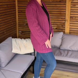 May include: A long, magenta-colored, open-front cardigan with a textured knit pattern. The cardigan is worn over a black top and paired with blue jeans and black boots. The outfit is worn in an outdoor setting.