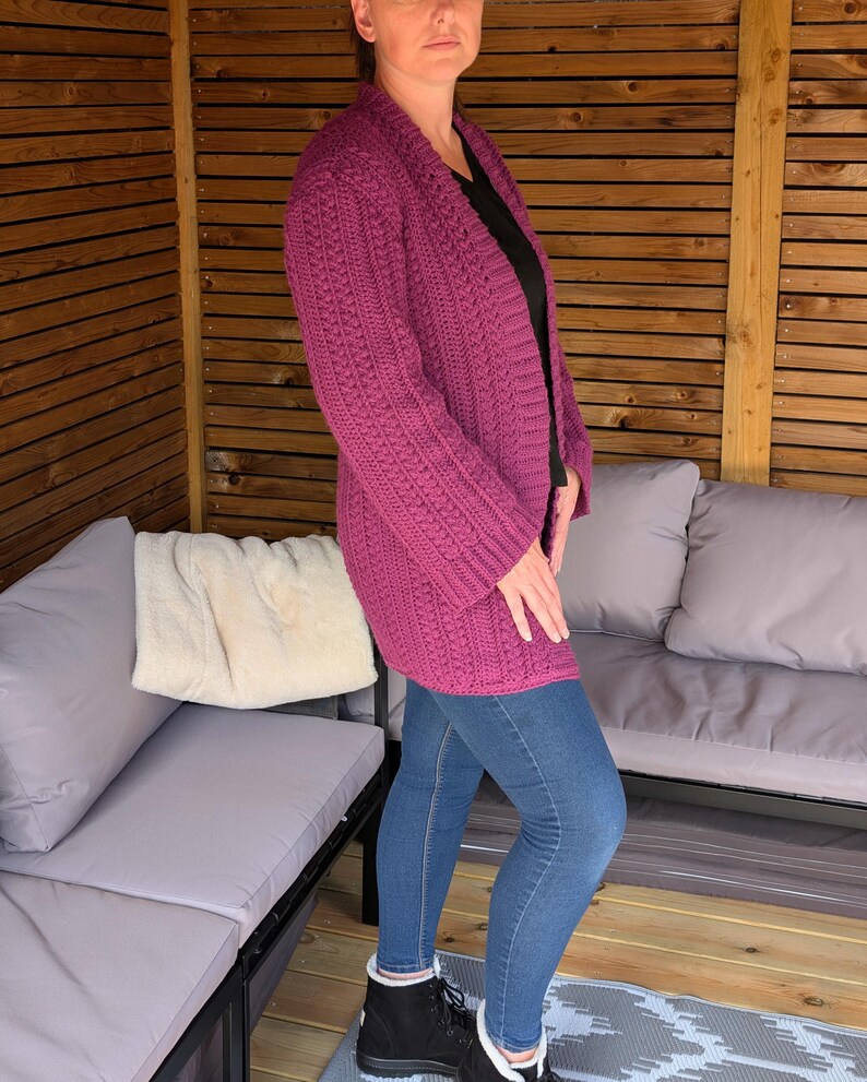 May include: A long, magenta-colored, open-front cardigan with a textured knit pattern. The cardigan is worn over a black top and paired with blue jeans and black boots. The outfit is worn in an outdoor setting.