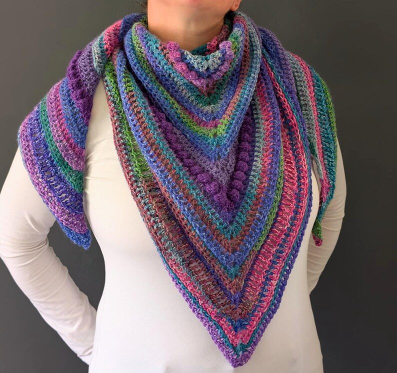 May include: A colorful, handmade crochet shawl in shades of purple, blue, green, and pink. The triangular shawl features a textured stitch pattern and is draped over the shoulders.