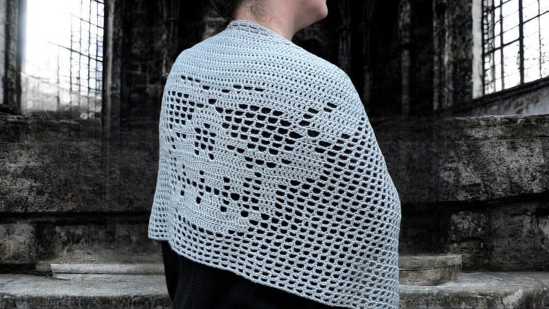 May include: A light blue crocheted shawl with a floral design. The shawl is draped over the shoulders, showcasing the intricate openwork pattern. The background is a dark, stone structure.
