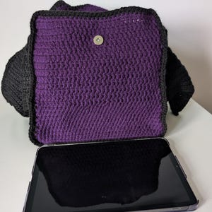 May include: A handmade crochet bag in purple and black. The bag features a rectangular shape with a flap closure and a magnetic snap. The bag is shown with a tablet in front of it. The bag is made of a thick yarn.