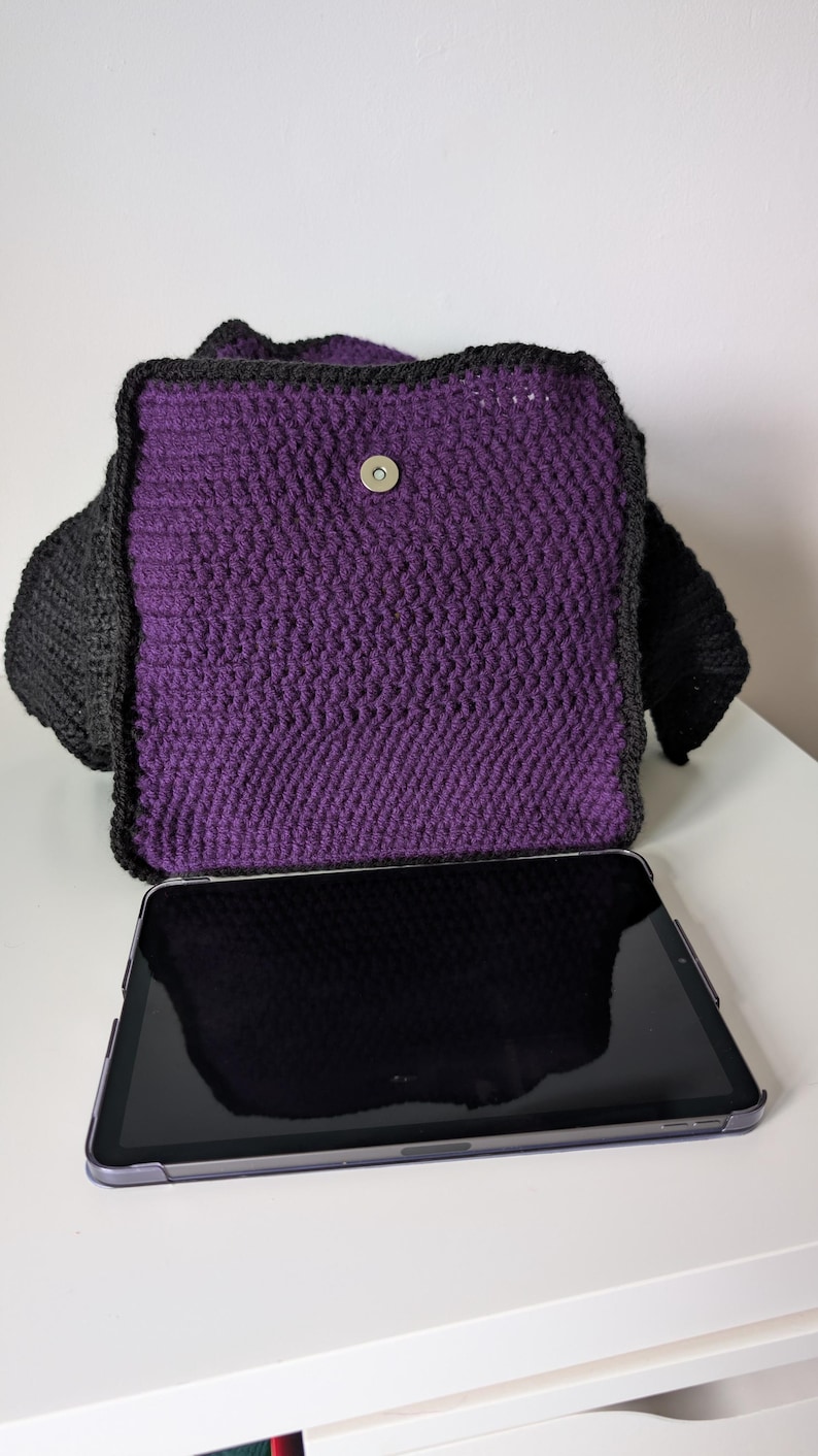 May include: A handmade crochet bag in purple and black. The bag features a rectangular shape with a flap closure and a magnetic snap. The bag is shown with a tablet in front of it. The bag is made of a thick yarn.