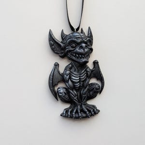 May include: A dark gray gargoyle pendant with a black cord. The pendant features a detailed design of a seated gargoyle with wings, horns, and a menacing expression. The gargoyle is approximately 3 inches tall.