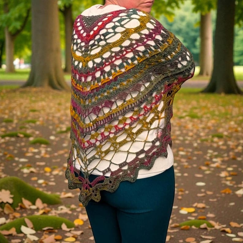 May include: A multi-coloured, open-weave shawl in shades of green, red, yellow, and brown. The shawl is draped over a person's shoulders, highlighting its intricate crochet design. The background shows a park with trees and fallen leaves.
