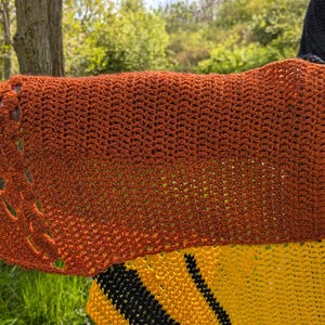 May include: A handmade crochet shawl in shades of orange and brown, with a textured stitch pattern and decorative cutouts along the edge. The shawl is part of a larger garment with yellow and black stripes, and a black hood.