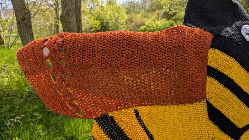 May include: A handmade crochet shawl in shades of orange and brown, with a textured stitch pattern and decorative cutouts along the edge. The shawl is part of a larger garment with yellow and black stripes, and a black hood.