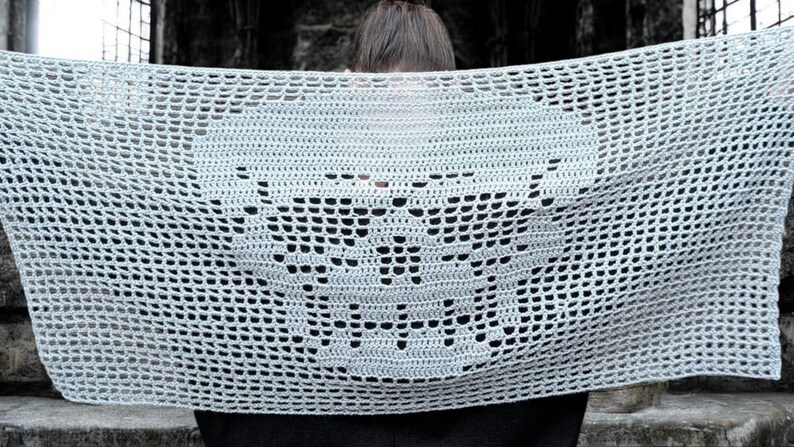 May include: A light gray crocheted shawl featuring a skull design. The open weave of the shawl creates a delicate, airy appearance. The skull is prominently displayed in the center of the shawl.