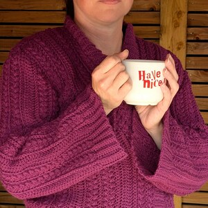 May include: A person wearing a magenta-colored, knitted robe with wide sleeves, holding a white mug. The mug has the words "Have nice" in red lettering. The robe has a textured, cable-knit design.