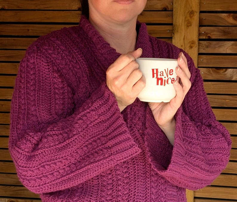 May include: A person wearing a magenta-colored, knitted robe with wide sleeves, holding a white mug. The mug has the words "Have nice" in red lettering. The robe has a textured, cable-knit design.
