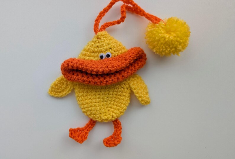 May include: A handmade yellow crochet duck ornament with an orange beak, feet, and a loop for hanging. The duck has googly eyes and a yellow pom-pom attached. This whimsical decoration is perfect for a nursery or children's room.