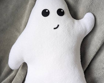 May include: A white plush ghost with black eyes and a smiling mouth. The ghost is lying on a gray blanket.