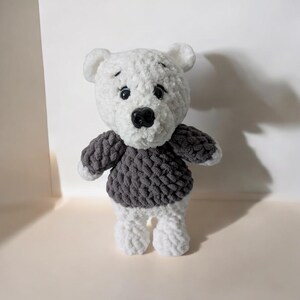 May include: A handmade white and gray crochet bear with a dark gray sweater. The bear has a black nose and small black eyes. The bear's arms and legs are white, and the sweater is a textured, chunky knit.