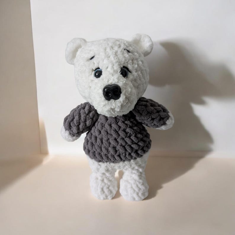 May include: A handmade white and gray crochet bear with a dark gray sweater. The bear has a black nose and small black eyes. The bear's arms and legs are white, and the sweater is a textured, chunky knit.