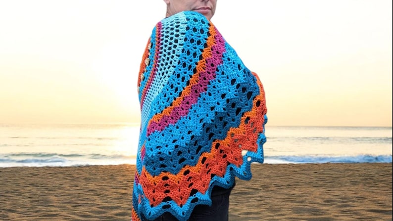 Op de afbeelding: Een kleurrijke gehaakte sjaal in blauw, oranje en roze tinten. De sjaal heeft een geschulpte rand en een geometrisch patroon. De achtergrond toont een strandsc&egrave;ne met de oceaan en een zonsondergang.