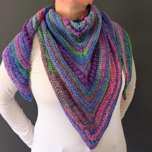 Pu&ograve; includere: Uno scialle all'uncinetto fatto a mano e colorato nei toni del viola, blu, verde e rosa. Lo scialle triangolare presenta un motivo a punto strutturato ed &egrave; drappeggiato sulle spalle.