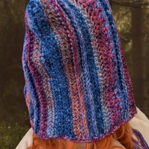 May include: A crocheted hooded hat with a striped pattern in shades of blue, purple, and pink. The hat has a long tassel hanging from the back.