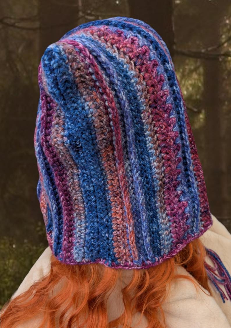 May include: A crocheted hooded hat with a striped pattern in shades of blue, purple, and pink. The hat has a long tassel hanging from the back.