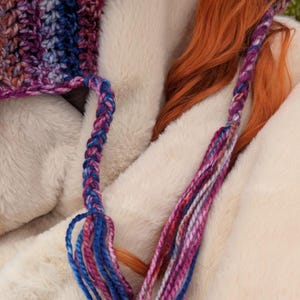 May include: A close-up of a crocheted hat with a braided strap and long, colorful tassels. The hat is made with a variety of shades of blue, pink, and purple yarn.