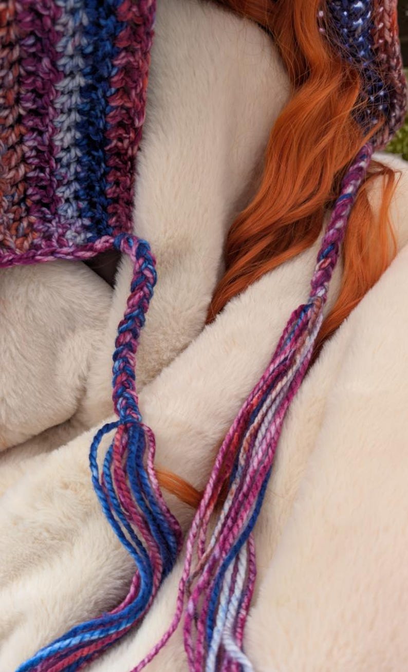 May include: A close-up of a crocheted hat with a braided strap and long, colorful tassels. The hat is made with a variety of shades of blue, pink, and purple yarn.