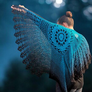 May include: A handmade crochet shawl in shades of blue and black. The shawl features a circular design on the back and a scalloped edge. The gradient color effect transitions from a bright blue to a dark black.
