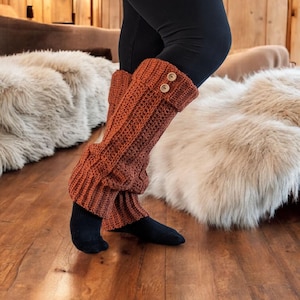May include: Rust-colored, chunky knit leg warmers with two wooden buttons at the top. The leg warmers are worn over black socks and leggings. The image is taken indoors on a wooden floor.
