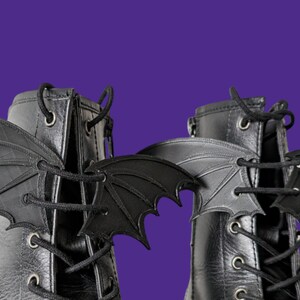 May include: A pair of black leather boots with black bat wings attached to the laces. The boots have a lace-up closure and a round toe.