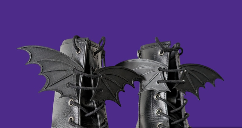 May include: A pair of black leather boots with black bat wings attached to the laces. The boots have a lace-up closure and a round toe.