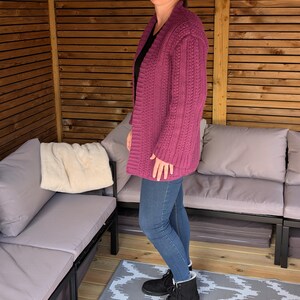 May include: A magenta-colored, open-front cardigan with a textured knit pattern. The long-sleeved sweater is worn over a black top and paired with blue jeans and black boots. The cardigan is a medium weight and appears to be handmade.