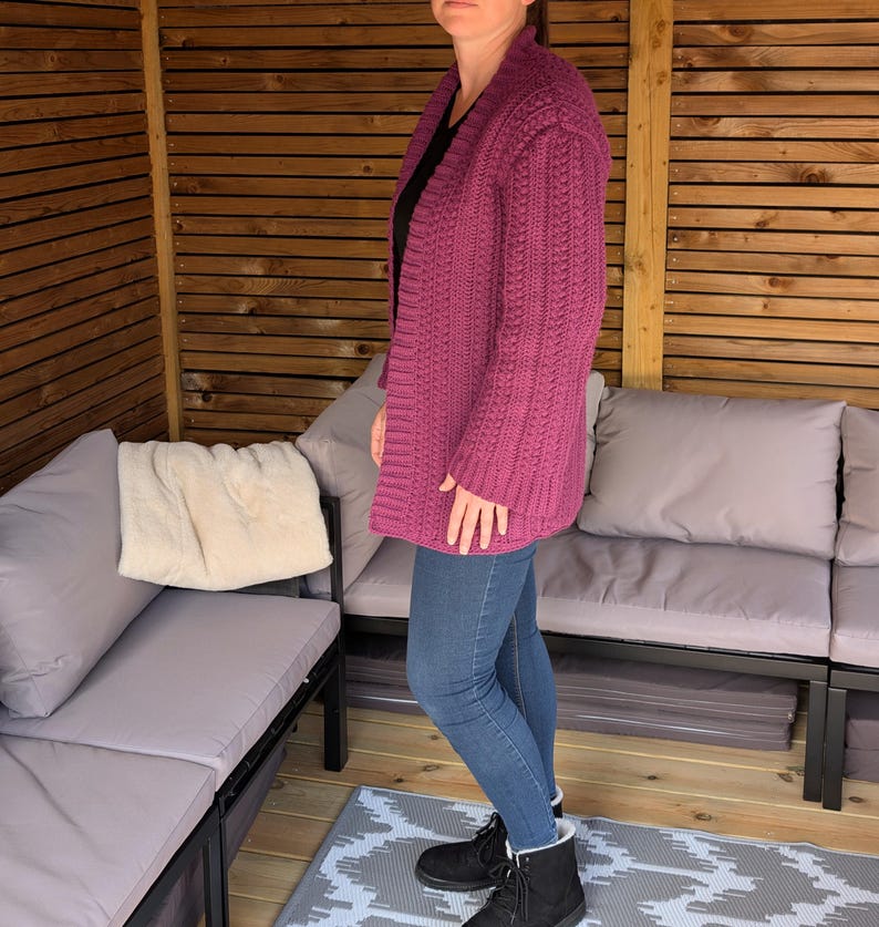 May include: A magenta-colored, open-front cardigan with a textured knit pattern. The long-sleeved sweater is worn over a black top and paired with blue jeans and black boots. The cardigan is a medium weight and appears to be handmade.