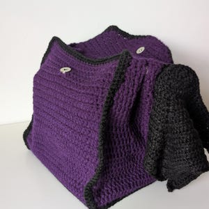 May include: A handmade, crocheted cowl in a deep purple with black trim and accents. The cowl has a textured stitch pattern and is secured with two small, round buttons. The black section appears to be a sleeve or extension of the cowl.