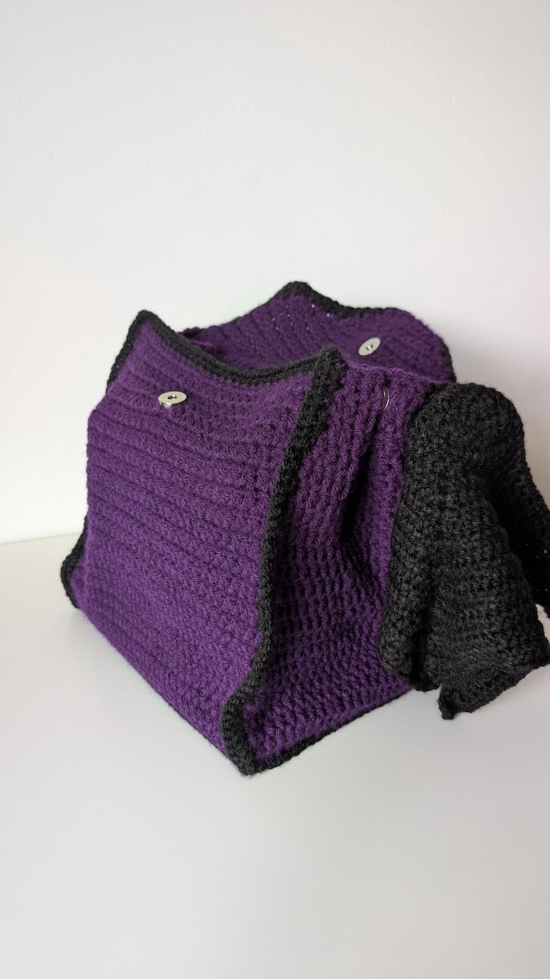 May include: A handmade, crocheted cowl in a deep purple with black trim and accents. The cowl has a textured stitch pattern and is secured with two small, round buttons. The black section appears to be a sleeve or extension of the cowl.