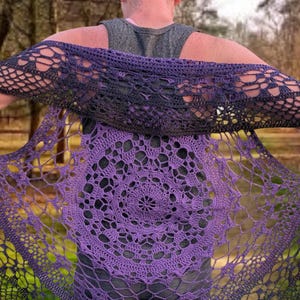 May include: A purple and black crochet shawl with a circular pattern. The shawl is draped over the shoulders, displaying the detailed openwork design. The colour transitions from deep purple to black, creating a gradient effect. The background is a blurred outdoor scene.