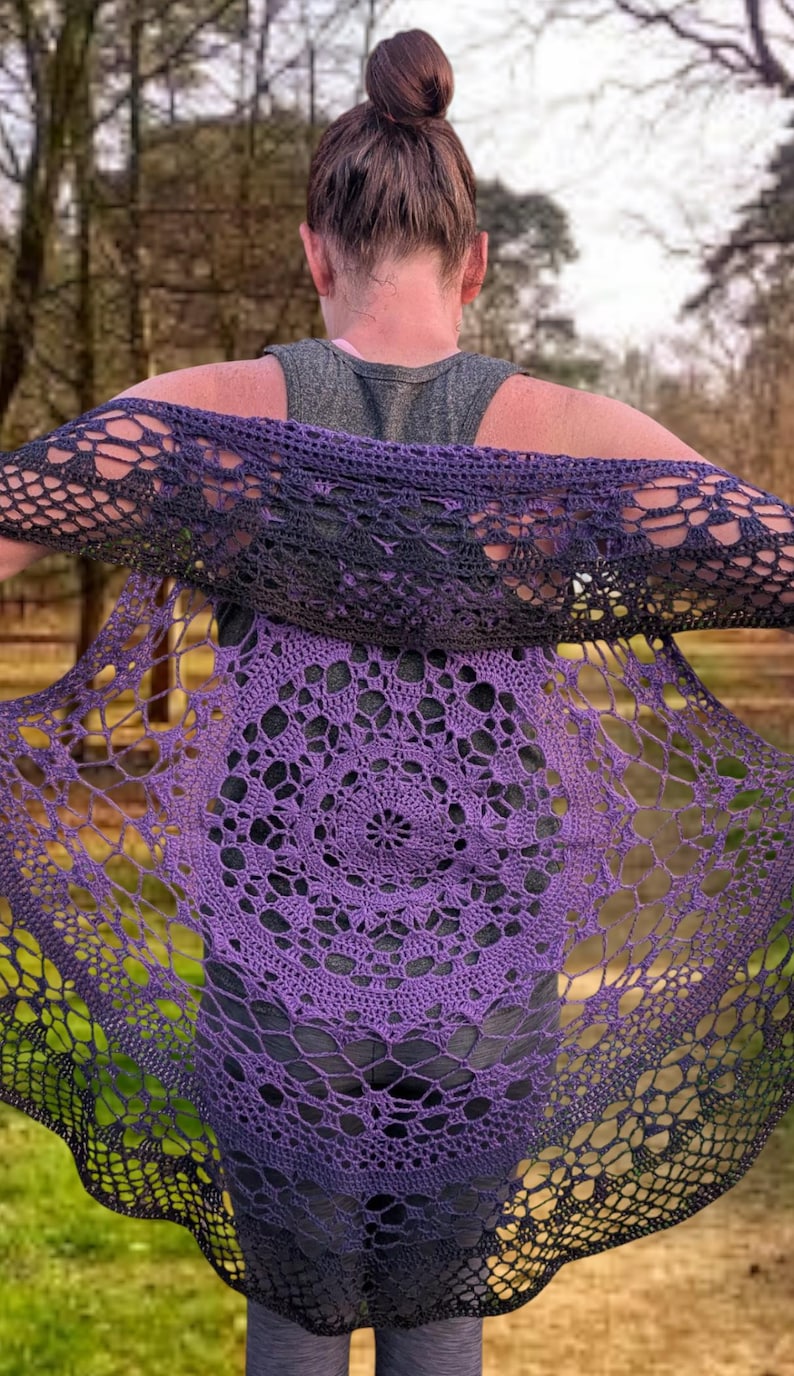 May include: A purple and black crochet shawl with a circular pattern. The shawl is draped over the shoulders, displaying the detailed openwork design. The colour transitions from deep purple to black, creating a gradient effect. The background is a blurred outdoor scene.