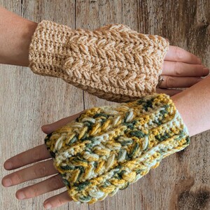 May include: Two handmade crochet fingerless gloves. One is a solid tan color, and the other is a mix of yellow, green, and white. The gloves have a textured, braided design. The gloves are displayed on a wooden surface.