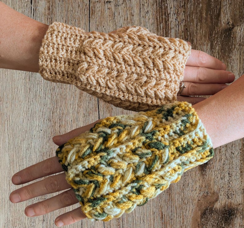 May include: Two handmade crochet fingerless gloves. One is a solid tan color, and the other is a mix of yellow, green, and white. The gloves have a textured, braided design. The gloves are displayed on a wooden surface.