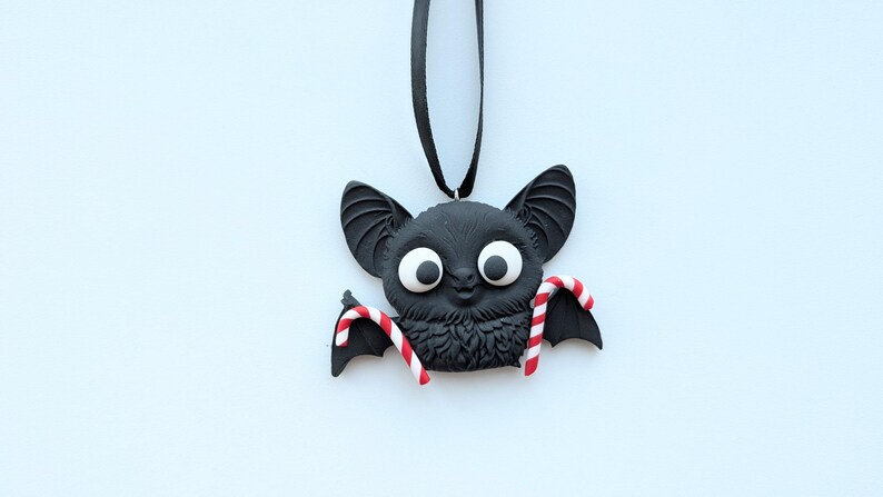 Puede incluir: Un adorno de murci&eacute;lago negro con ojos blancos grandes y bastones de caramelo rojos y blancos. El murci&eacute;lago tiene orejas grandes y cuelga de una cinta negra. El adorno probablemente est&aacute; hecho de arcilla.