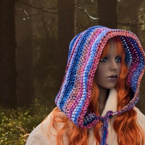 May include: A woman with long, red hair wears a colorful crocheted hooded scarf. The scarf is striped with shades of blue, pink, and purple. The woman is standing in a forest setting.