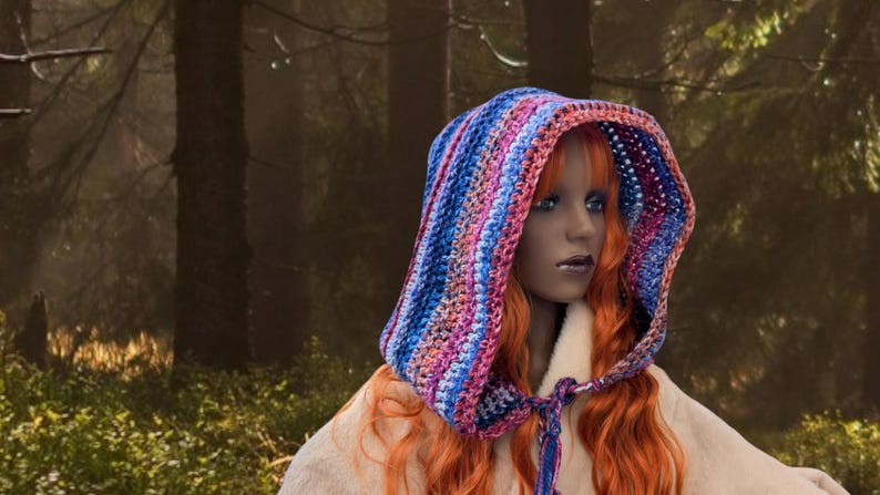 May include: A woman with long, red hair wears a colorful crocheted hooded scarf. The scarf is striped with shades of blue, pink, and purple. The woman is standing in a forest setting.