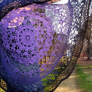 May include: A hand-crocheted shawl in a gradient of colours, shifting from deep purple to black. The shawl has an open, lacy design with a circular pattern in the centre. The shawl is being held up, displaying its intricate details.