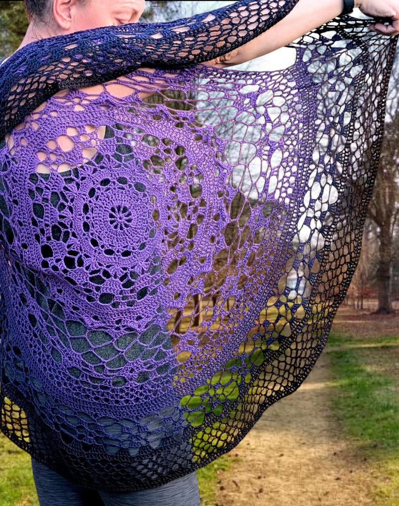 May include: A hand-crocheted shawl in a gradient of colours, shifting from deep purple to black. The shawl has an open, lacy design with a circular pattern in the centre. The shawl is being held up, displaying its intricate details.