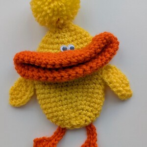 May include: A handmade, yellow crochet duck with an orange beak and feet. The duck has a yellow pom-pom on its head and googly eyes. The duck is a whimsical, handcrafted item.