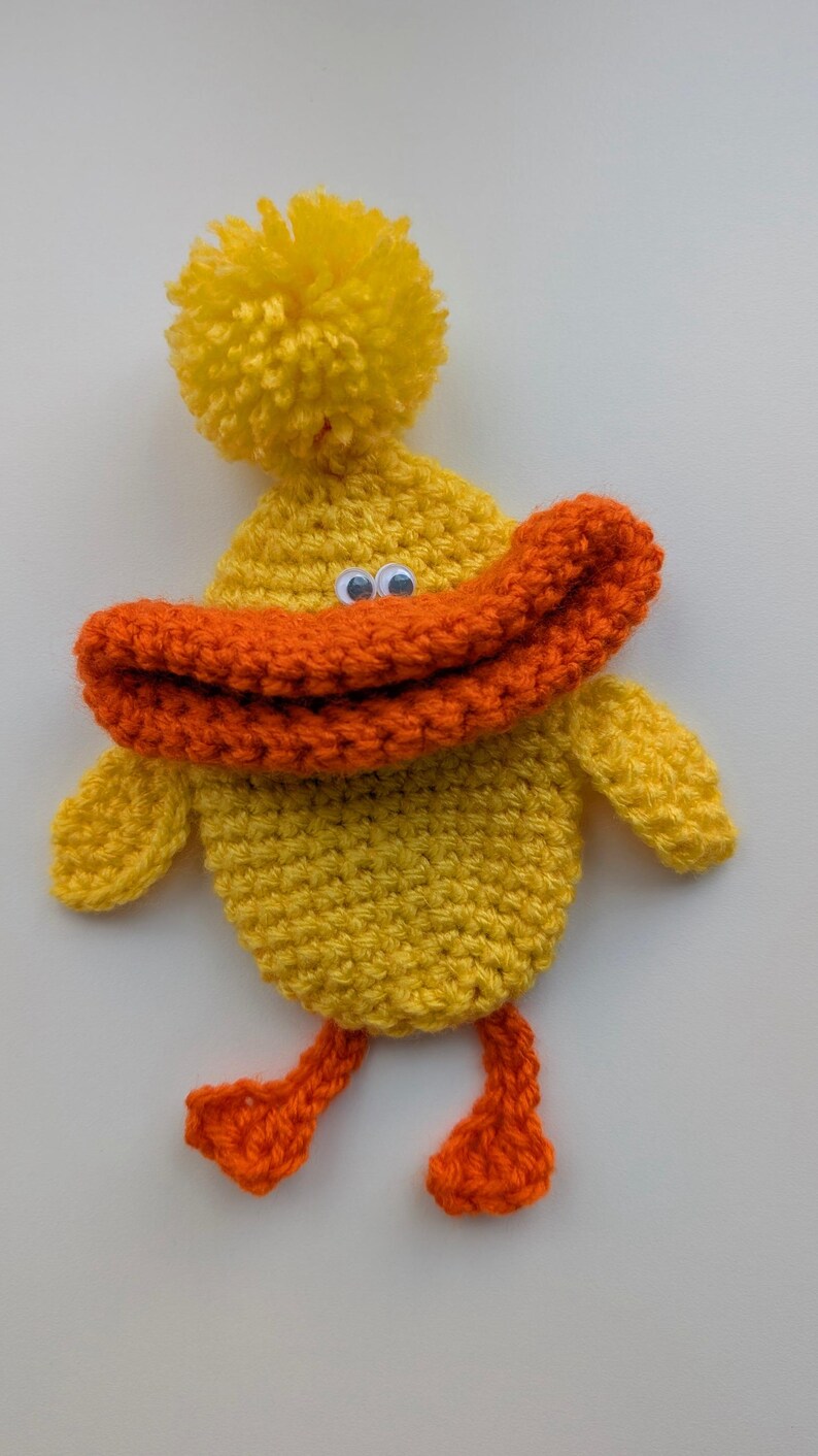 May include: A handmade, yellow crochet duck with an orange beak and feet. The duck has a yellow pom-pom on its head and googly eyes. The duck is a whimsical, handcrafted item.
