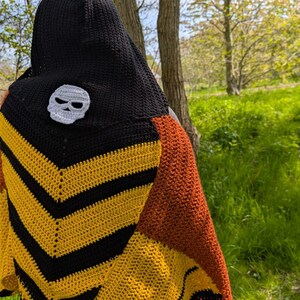 May include: A handmade crochet shawl with a black hood and a white skull applique. The shawl features a chevron pattern in yellow and black, with a section of orange-brown. The background is a natural outdoor setting with trees and green grass.