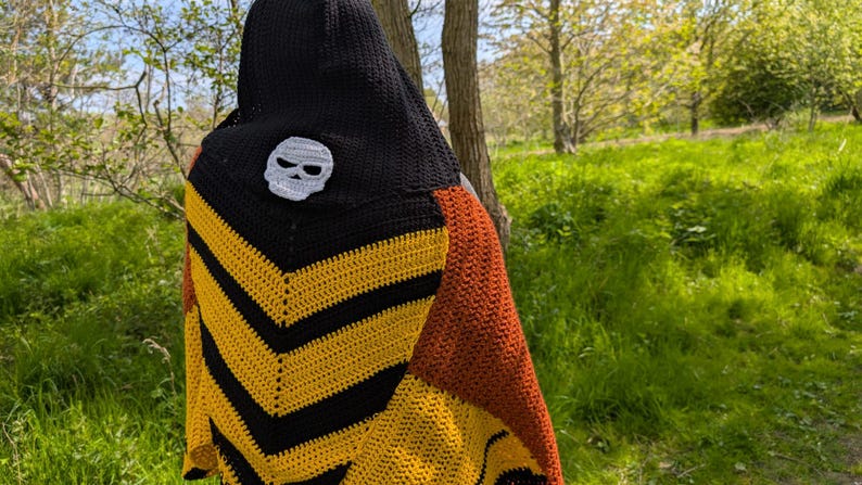 May include: A handmade crochet shawl with a black hood and a white skull applique. The shawl features a chevron pattern in yellow and black, with a section of orange-brown. The background is a natural outdoor setting with trees and green grass.