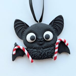 Puede incluir: Un adorno negro con forma de murci&eacute;lago con ojos blancos grandes y dos bastones de caramelo. El murci&eacute;lago tiene orejas grandes y un pecho peludo. Los bastones de caramelo son de rayas rojas y blancas. El adorno cuelga de una cinta negra.