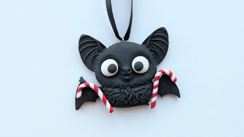 Puede incluir: Un adorno negro con forma de murci&eacute;lago con ojos blancos grandes y dos bastones de caramelo. El murci&eacute;lago tiene orejas grandes y un pecho peludo. Los bastones de caramelo son de rayas rojas y blancas. El adorno cuelga de una cinta negra.